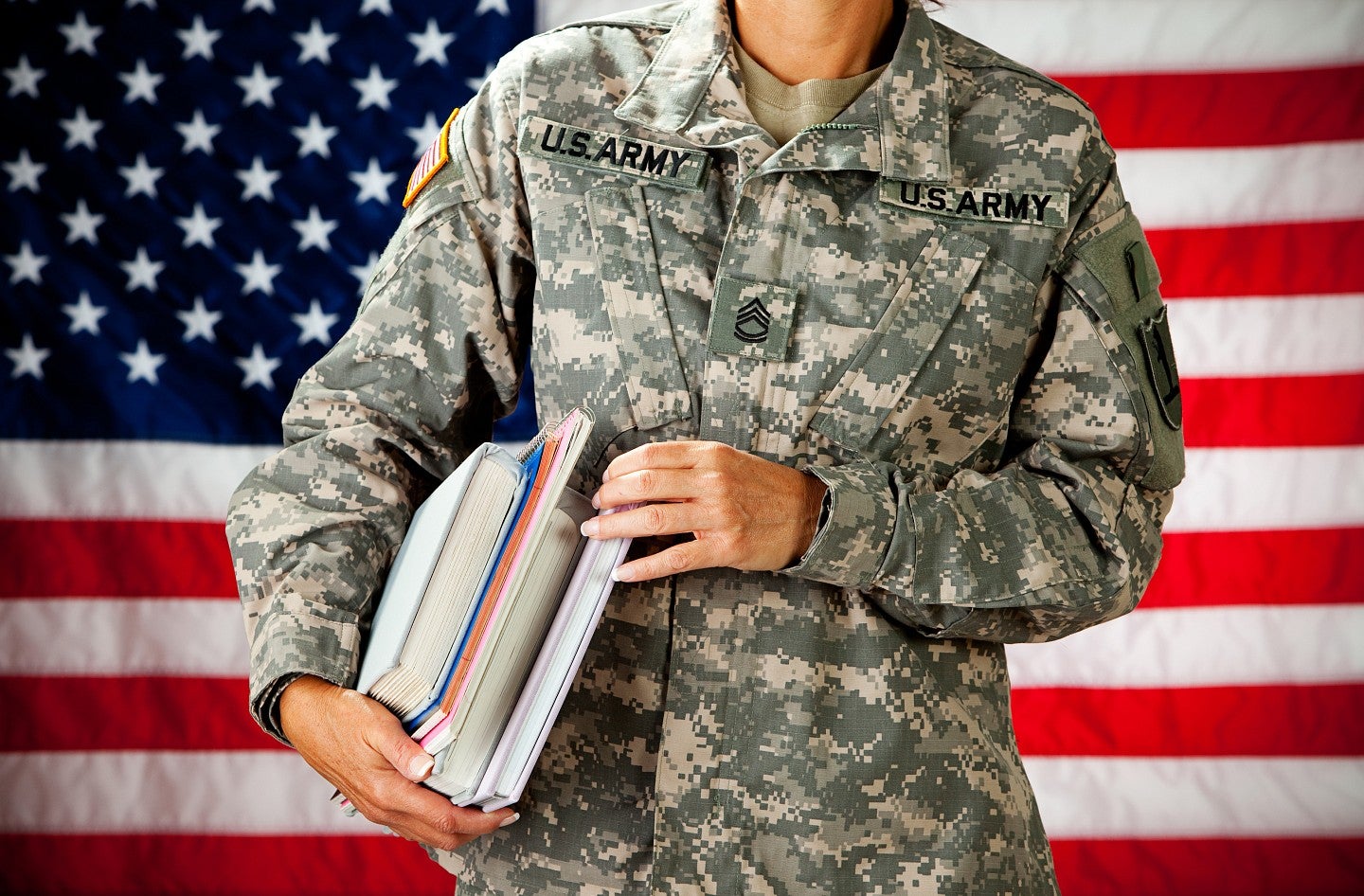 military person holding textbooks standing in front of american flag