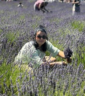 A woman wearing a light blue shirt and sunglasses smiling at the camera in a field of lavender.